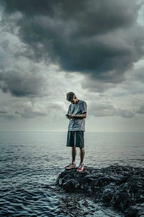 man in white long sleeve shirt and blue shorts standing on rock in front of sea using a phone